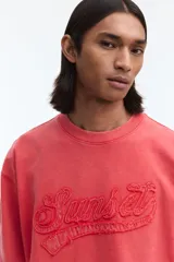 Remera boxy de algodón liviano en color coral, con efecto acid wash, hombros caídos, cuello redondo ribeteado y corte oversized. Cuenta con bordado en relieve con la palabra 'Sunset' en el frente.
