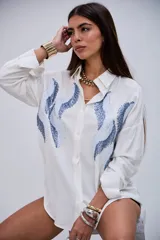 Camisa blanca de manga larga con cuello clásico y botones frontales. Presenta un diseño de ondas en tonos azules con lentejuelas y pequeños apliques metálicos en el frente.