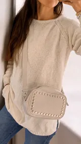Cartera bandolera de cuero sintético color beige, con diseño rectangular y detalle de tachas metálicas circulares en el contorno frontal. Cuenta con correa larga ajustable y desmontable mediante mosquetones, cierre superior y bolsillos internos y externos.