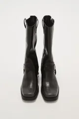 Bota de caña media estilo western, confeccionada en cuero sintético negro. Presenta puntera cuadrada, taco bajo cuadrado y una correa decorativa con tachas metálicas y argolla en el tobillo.