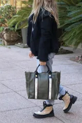 Bolso tipo shopper color verde oliva, con doble correa de mano en cuero negro y detalles de cintas verticales con logo estampado. Cuenta con cierre superior, bolsillos laterales y un diseño espacioso ideal para uso diario.