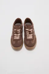 Zapatillas urbanas de corte bajo en tonos beige y tostado, combinando paneles de cuerina y gamuza sintética. Cuentan con cordones anchos tipo cinta de raso en color crudo, pespuntes decorativos y suela de goma al tono.