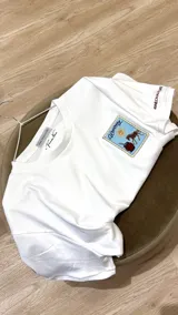 Remera de algodón premium color crema, con corte holgado y cuello redondo. Presenta un bordado en el pecho con diseño de estampilla que incluye un sol, un mate y un caballo, además de un bordado de herradura en la manga derecha.