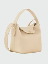 Cartera bucket S Longchamp modelo Le Cadence, color beige, confeccionada en cuero graneado. Cuenta con un asa superior fija, solapa frontal con botón a presión dorado y una correa ajustable para llevar cruzada o al hombro.