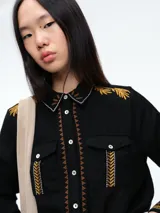 Camisa de color crudo con detalles de bordados en tonos mostaza y marrón en el cuello, hombros, bolsillos frontales y puños. Presenta cierre frontal con botones y dos bolsillos tipo cargo en el pecho.