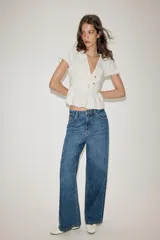 Top de color blanco con escote en pico, mangas cortas abullonadas y bajo con diseño peplum. Presenta detalle de botones dorados en la parte frontal y cierre lateral con cremallera oculta.
