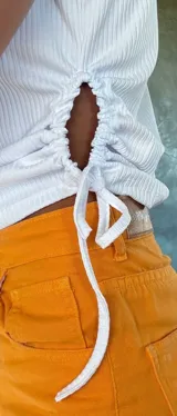 Top sin mangas de cuello alto, confeccionado en tejido canalé blanco. Presenta un diseño entallado con aberturas laterales ajustables mediante cordones.