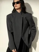 Blazer sastrero de corte oversized en color gris oscuro, con hombros estructurados, cuello mao y cierre frontal cruzado con un botón. Presenta bolsillos laterales con solapa y un bolsillo superior en el pecho.