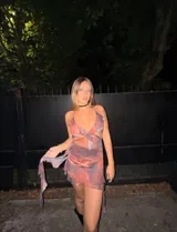 Vestido corto con estampado tie-dye en tonos naranja, azul y blanco. Presenta escote halter con volados, aberturas laterales en la cintura y falda fruncida con tiras largas.