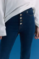 Jean de tiro alto con corte oxford, confeccionado en denim azul oscuro. Presenta cierre frontal con cinco botones metálicos visibles y aberturas en el bajo de las piernas.