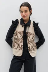 Chaleco de lana color beige con bordados florales artesanales en tonos marrones y blancos. Presenta un diseño sin mangas con volados en los hombros y cierre frontal mediante lazos negros ajustables.