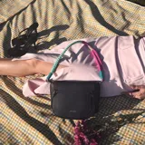 Cartera tipo bandolera negra de formato compacto, con solapa frontal, líneas curvas y correa de hombro tejida multicolor en tonos turquesa y fucsia.