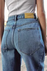 Jeans de tiro alto con diseño barrel, confeccionados en denim azul. Presentan cinco bolsillos, cierre frontal con botones y detalle de costuras verticales en el frente y la espalda. Su corte es holgado en la pernera y se estrecha hacia el tobillo.