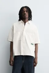 Camisa de algodón con estructura canvas, color rojo lavado, de corte relajado (relaxed fit) y manga corta. Posee cuello solapa, dos bolsillos de plastrón en el pecho y cierre frontal de botonadura. Presenta bordado y estampado de texto a contraste en el delantero.