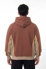 Hoodie oversize color marrón con paneles laterales en beige y detalles de costuras en contraste amarillo. Presenta capucha con cordones ajustables, bolsillo tipo canguro frontal y logo bordado en el pecho.