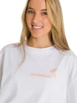 Remera blanca de corte oversize con cuello redondo y mangas cortas. Presenta un estampado gráfico en el pecho con el logo de la marca y una palmera en color salmón.