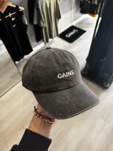 Gorra con visera de efecto acid wash en color gris oscuro, con diseño de seis paneles y el texto 'GAINS.' bordado en blanco en el frente.