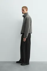 Pantalón negro de corte relaxed fit y estilo wide leg, con pliegues frontales dobles, bolsillos delanteros y detalle de bolsillos traseros de vivo. Cierre frontal cruzado con cremallera y gancho metálico oculto.