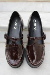 Zapato tipo guillermina en cuero charol color bordó, con suela track negra de goma, cierre con hebilla ajustable y detalle de herrajes metálicos dorados.