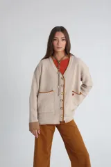 Saco de punto color beige con textura acanalada, cuello en V y cierre frontal con botones. Presenta bolsillos delanteros con bordes en color marrón y un pequeño detalle bordado en rojo sobre uno de los bolsillos.