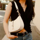 Cartera tipo hobo de la marca Guess, color beige con detalles en negro. Presenta un diseño compacto con cierre superior, correa de hombro con eslabones metálicos dorados y un detalle central con el logo de la marca en dorado.