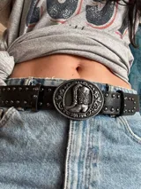 Cinturón negro de cuero sintético con hebilla plateada con diseño de botas cowboy.