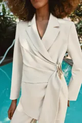 Blazer de corte estructurado en color beige, con cuello solapa y manga larga. Presenta un diseño cruzado con cierre mediante lazada del mismo tejido y un distintivo aplique metálico dorado en el frente.
