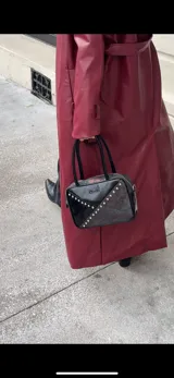 Cartera rígida negra con detalles en rojo y tachas plateadas. Tiene herrajes y cierre metálico plateado marca YKK, asa fina para llevar al hombro o en la mano y correa larga extra regulable. El interior es forrado, tiene un bolsillo con cierre enmarcado en cuero, y un segundo bolsillo plaqué.