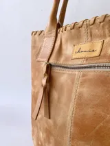 Cartera tipo tote de cuero color camel, con diseño artesanal que presenta costuras decorativas visibles en los laterales y el frente. Cuenta con doble asa de hombro y cierre superior.