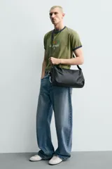 Bolso tipo bandolera de diseño minimalista en color negro, con cierre de cremallera superior y asa de hombro ajustable. Incluye un detalle decorativo de charms metálicos en uno de los laterales.