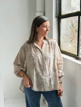 Camisa de lino color beige con diseño oversize, cuello camisero mao, cierre frontal con botones ocultos, mangas largas abullonadas con puños elásticos y delicados bordados florales tono sobre tono en el frente y las mangas.