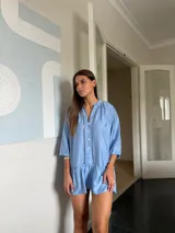 Vestido corto azul claro de manga larga, con cuello mao y cierre de botones frontal. Presenta detalles de encaje o bordado en las costuras verticales del frente y en la zona de los hombros. La manga se ajusta con lazo y el ruedo tiene un volante fruncido.