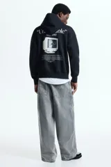 Canguro de corte oversized en mezcla de algodón, color negro, con capucha de doble capa, hombros caídos y bolsillo frontal tipo canguro. Presenta un estampado tipográfico pequeño en el pecho y puños y bajo acanalados.