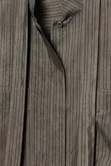 Blusa color gris con finas rayas verticales negras, cuello redondo con lazo del mismo tejido, manga larga con puño, bolsillos de parche en el pecho, bajo asimétrico y cierre frontal oculto con botones.