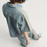 Camisa de jean de manga corta Adidas, confeccionada en 100% algodón, con diseño inspirado en la ropa de trabajo, cierre frontal con broches a presión, cuello camisero, un bolsillo en el pecho con solapa y las clásicas tres tiras de la marca en las mangas.