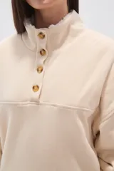 Buzo de algodón color tostado, con cuello polo y cierre frontal parcial con botones. Presenta un detalle de forro de corderito sintético blanco en el cuello y mangas largas con diseño holgado.