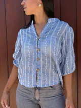 Camisa de corte recto y calce holgado, confeccionada en tela liviana color celeste con estampado de rayas verticales blancas con detalles de hojas. Presenta cuello en V, cierre frontal con botones de madera y mangas tres cuartos con terminación elástica.