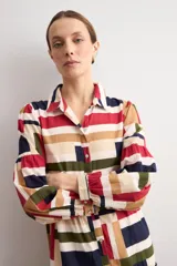 Vestido largo tipo camisero con estampado geométrico multicolor de bloques en tonos rojo, azul, verde y tostado. Presenta cuello camisero, cierre frontal con botonadura completa, mangas largas abullonadas y diseño escalonado en la falda.