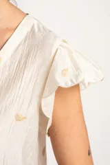 Blusa blanca sin mangas con escote en V, cierre frontal con botones y mangas con volados. Presenta pequeños bordados sutiles distribuidos por toda la prenda.