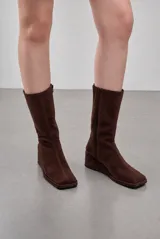 Bota de caña alta confeccionada en gamuza color marrón, con punta cuadrada, taco cuña corrido forrado y cierre lateral.