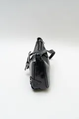 Bolso de hombro tipo baguette en color negro con acabado brillante. Presenta un diseño estructurado con una hebilla decorativa metálica en el frente y detalles de tachas plateadas en los laterales.