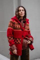 Saco tejido a mano con diseño jacquard en tonos rojo, naranja y marrón. Presenta cuello tipo polo, cierre frontal con botones grandes, mangas largas y tajos laterales en el ruedo.