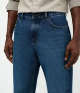Pantalón de jean de corte recto en denim de algodón con lavado oscuro uniforme, cintura con presillas, cierre frontal con botón y bolsillos funcionales.