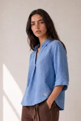 Blusa azul de lino con cuello camisero, escote en V y mangas tres cuartos con vuelta. Presenta un diseño holgado con dobladillo redondeado y un pequeño parche con logo bordado en el lateral inferior.