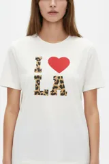 Remera blanca de manga corta y cuello redondo, con corte holgado y estampado frontal que dice 'I <3 LA' con letras de animal print y un corazón rojo.