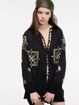 Camisa negra de manga larga con cuello camisero y bordados geométricos en hilo dorado. Presenta un borde en zigzag dorado en la abertura frontal y un largo irregular en la espalda.