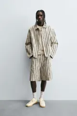 Bermuda de hombre color beige con estampado de rayas verticales azules con efecto desgastado. Corte relaxed fit, con cierre frontal de cremallera y botón, bolsillos delanteros y bolsillos traseros de plastrón.