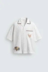 Camisa de corte relajado confeccionada en algodón, con cuello bowling y manga corta. Presenta un bolsillo en el pecho con vivo a contraste y bordados decorativos en el frente. Cierre frontal mediante botones.