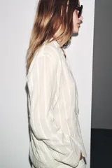 Camisa de algodón color crema o blanco roto, con textura de rayas verticales sutiles. Presenta cuello solapa, escote pico y manga larga.