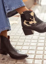 Botas texanas de caña baja confeccionadas en cuero natural. Presentan un diseño combinado con cuero liso negro y detalle de cuero con estampado de vaca en el talón. Cuentan con taco cuadrado de 4,5 cm, cierre lateral y puntera estilizada con costuras decorativas.